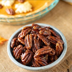 Könnte beinhalten: Nahaufnahme einer Metallschüssel gefüllt mit glasierten Pekannusshälften. Die Pekannüsse haben eine satte braune Farbe und wirken glänzend. Im Hintergrund ist ein Kürbiskuchen zu sehen, was auf ein Herbst- oder Feiertags-Thema hindeutet.