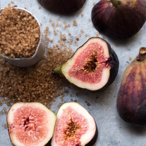 May include: An overhead view of fresh figs and brown sugar. Some figs are whole, others are cut open, showing a vibrant pink interior. A small metal container holds brown sugar, scattered around the figs.