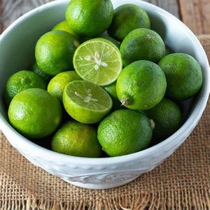 May include: A white bowl filled with fresh, green limes, some halved to reveal the juicy interior. The bowl sits on a burlap cloth over a wooden surface, creating a rustic and natural aesthetic. The limes are a vibrant green.