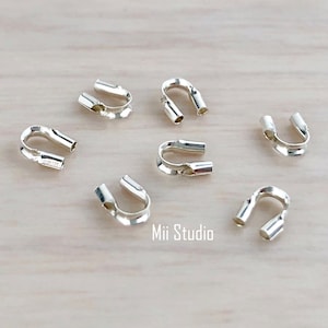 Könnte beinhalten: Sieben silberfarbene Metall-Quetschperlen, auch bekannt als Quetschhülsen, liegen auf einer hellbraunen Holzoberfläche. Die Perlen werden verwendet, um Schmuckteile zu sichern und ein professionelles Finish zu erzielen.