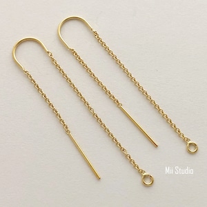 May include: A pair of gold dangle earrings with a simple curved top and a chain that extends down to a small circle. The earrings are shown on a white background.