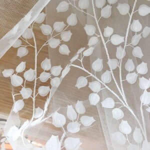 May include: White embroidered tulle fabric with a floral pattern. The design features raised, off-white leaves and blossoms on a sheer, white mesh background.  The fabric is delicate and lightweight, suitable for apparel or home decor projects. The floral motif is repeated consistently across the material.