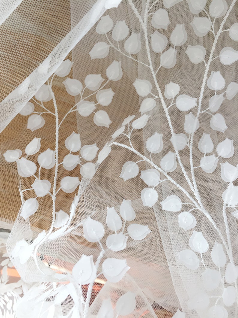May include: White embroidered tulle fabric with a floral pattern. The design features raised, off-white leaves and blossoms on a sheer, white mesh background.  The fabric is delicate and lightweight, suitable for apparel or home decor projects. The floral motif is repeated consistently across the material.