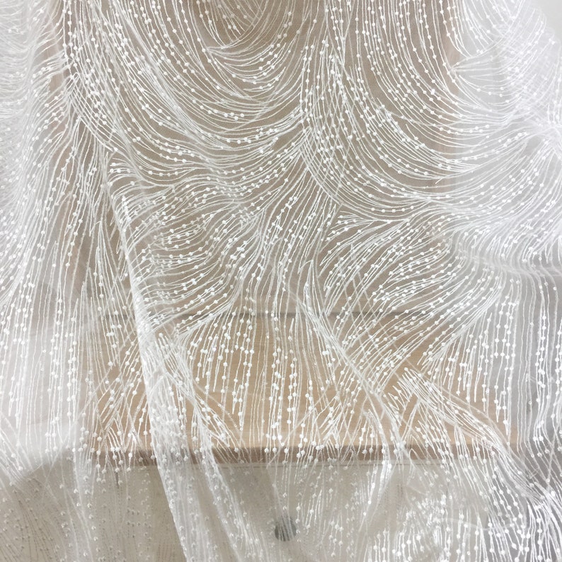 Peut inclure: Tissu de dentelle blanche perl&eacute;e avec un motif ondul&eacute;. Le tissu est transparent et d&eacute;licat, avec de petites perles blanches &eacute;parpill&eacute;es.