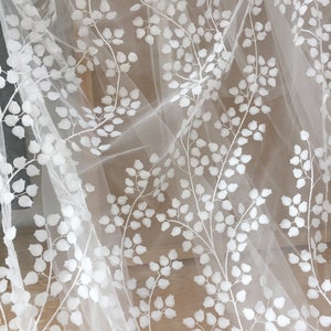 May include: White embroidered tulle fabric with a delicate floral pattern. The design features small, white leaves and branches on a sheer, white background.  This elegant fabric is perfect for wedding apparel, bridal veils, or other special occasion garments. The texture is soft and lightweight, ideal for creating flowing and romantic designs.