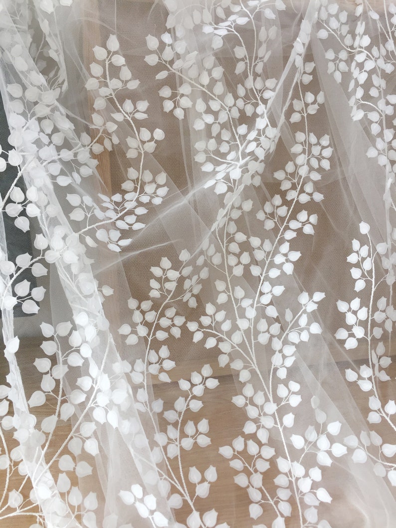May include: White embroidered tulle fabric with a delicate floral pattern. The design features small, white leaves and branches on a sheer, white background.  This elegant fabric is perfect for wedding apparel, bridal veils, or other special occasion garments. The texture is soft and lightweight, ideal for creating flowing and romantic designs.