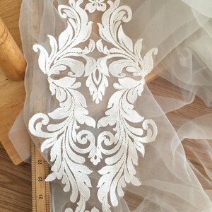 Puede incluir: Aplique de encaje blanco con un dise&ntilde;o floral. El aplique est&aacute; sobre una tela blanca transparente.