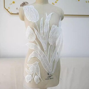 May include: White floral lace applique featuring a tulip design. The applique is made of delicate lace with intricate detailing, including the petals, leaves, and stems. The design is displayed on a mannequin, showcasing the potential for garment embellishment.