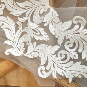 Puede incluir: Borde de encaje blanco bordado con un delicado patr&oacute;n floral sobre una tela de malla transparente.