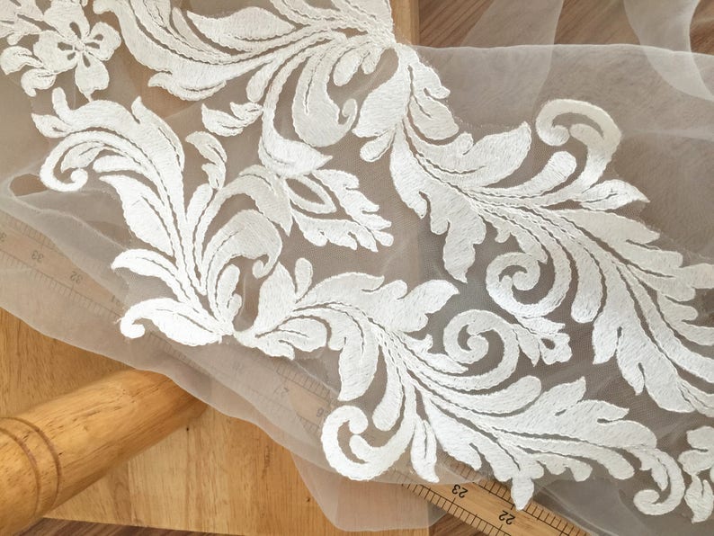 Puede incluir: Borde de encaje blanco bordado con un delicado patr&oacute;n floral sobre una tela de malla transparente.