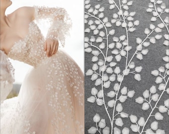 May include: A strapless ivory wedding dress with floral embellishments and long sleeves. The dress features a full skirt and is adorned with delicate, three-dimensional floral appliques. A close-up shows white leaf-like embroidery on a gray background.