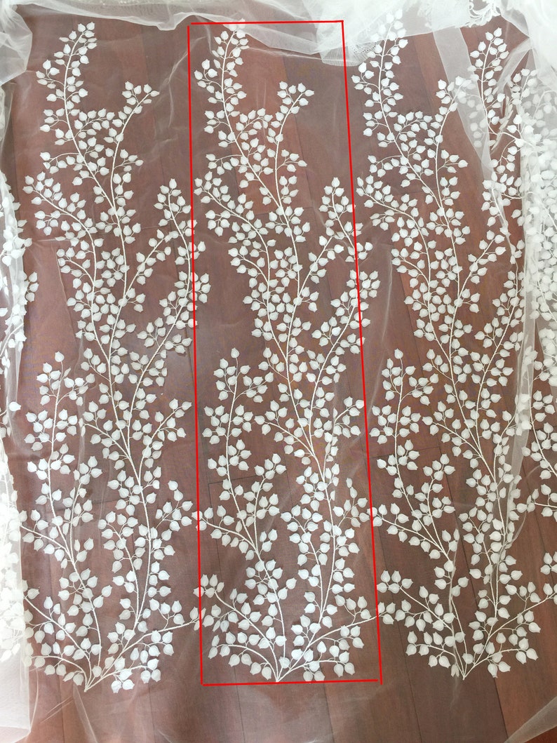 May include: White embroidered floral lace fabric on a sheer mesh background. The design features repeating patterns of delicate white leaves and branches, creating an elegant and intricate look. The lace is shown in a rectangular section, highlighting the detail and texture of the embroidery. This high-quality fabric is perfect for wedding dresses, bridal gowns, veils, and other special occasion garments.