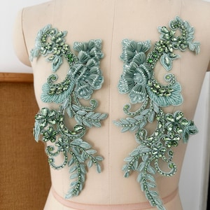 Puede incluir: Dos apliques florales a juego en tonos verdes y turquesas. Cada aplique presenta hojas bordadas, detalles de cuentas y detalles de flores de tela. Los apliques se muestran en un maniquí.