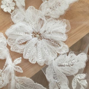 May include: White lace fabric with floral embroidery and sequins. The lace features a delicate floral pattern with a large, central flower.