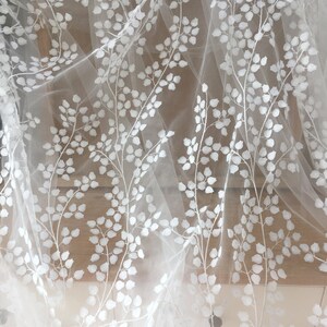 May include: White embroidered tulle fabric features a delicate floral pattern. The design showcases repeating branches with small, light-colored leaves, creating an elegant and airy aesthetic.  This sheer, lightweight textile is ideal for various applications, such as wedding apparel, home decor, and crafting projects. The embroidered details add a touch of sophistication and texture to the fabric.