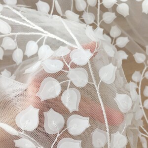 May include: White embroidered tulle fabric with a floral design. The fabric features delicate, raised white flowers on a sheer white background.  The flowers are arranged in a repeating pattern along elegant stems, creating a lovely, romantic aesthetic. This high-quality fabric is perfect for wedding apparel, bridal gowns, or other special occasion garments.