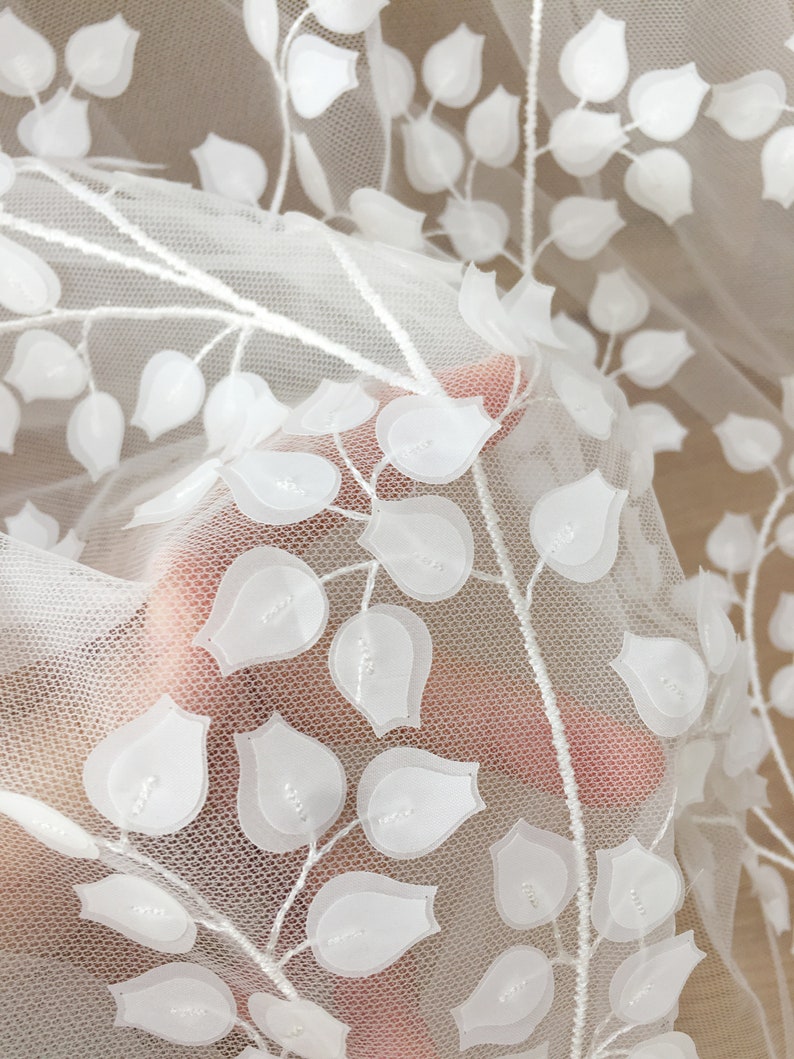 May include: White embroidered tulle fabric with a floral design. The fabric features delicate, raised white flowers on a sheer white background.  The flowers are arranged in a repeating pattern along elegant stems, creating a lovely, romantic aesthetic. This high-quality fabric is perfect for wedding apparel, bridal gowns, or other special occasion garments.