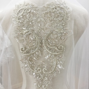 May include: A pair of silver beaded and sequined appliques in a paisley design. The appliques are on a white sheer fabric background.