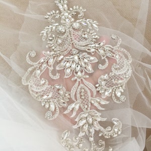May include: A delicate white lace applique with intricate floral designs and sparkling crystal embellishments. The applique is perfect for adding a touch of elegance to wedding dresses, bridal accessories, or other special occasion garments.