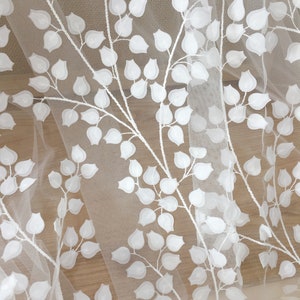 May include: White embroidered tulle fabric with a leaf pattern. The leaves are raised and textured, creating a three-dimensional effect. The fabric is sheer and delicate, with a soft, flowing drape.  This elegant fabric is ideal for wedding dresses, bridal veils, and other special occasion garments. The design features repeating branches with small, rounded leaves, creating a botanical motif. The white embroidery stands out against the sheer tulle background.