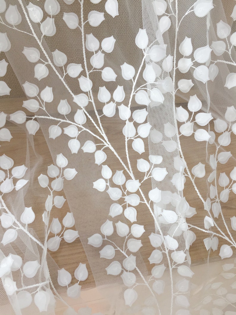 May include: White embroidered tulle fabric with a leaf pattern. The leaves are raised and textured, creating a three-dimensional effect. The fabric is sheer and delicate, with a soft, flowing drape.  This elegant fabric is ideal for wedding dresses, bridal veils, and other special occasion garments. The design features repeating branches with small, rounded leaves, creating a botanical motif. The white embroidery stands out against the sheer tulle background.
