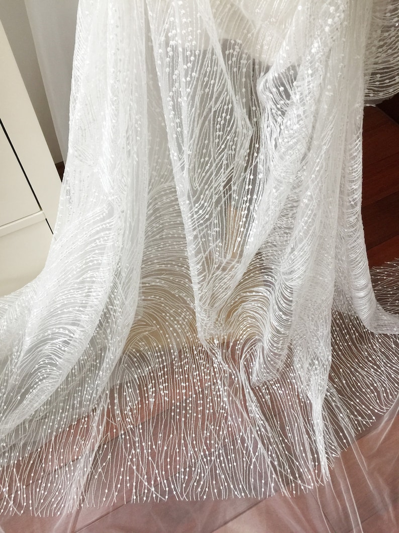 Peut inclure: Tulle blanc avec un motif d&eacute;licat de petites perles blanches. Le tissu est transparent et a une texture douce et fluide.