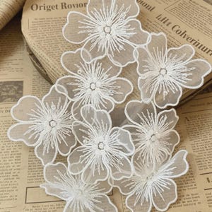 May include: Close-up of several white, embroidered flower appliques. Each flower has delicate, detailed stitching and a sheer, layered appearance. The appliques are arranged on a vintage newspaper background, creating a contrast between the delicate flowers and the aged paper.