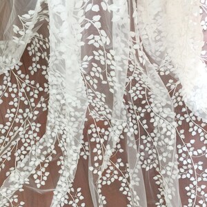 May include: Ivory embroidered tulle fabric with a delicate floral pattern. The design features white leaves and blossoms on a sheer, lightweight tulle background.  This elegant fabric is ideal for wedding dresses, veils, or other special occasion garments. The intricate embroidery adds texture and dimension to the fabric, creating a luxurious and romantic look.