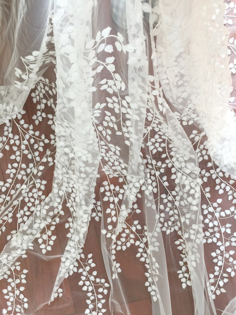 May include: Ivory embroidered tulle fabric with a delicate floral pattern. The design features white leaves and blossoms on a sheer, lightweight tulle background.  This elegant fabric is ideal for wedding dresses, veils, or other special occasion garments. The intricate embroidery adds texture and dimension to the fabric, creating a luxurious and romantic look.