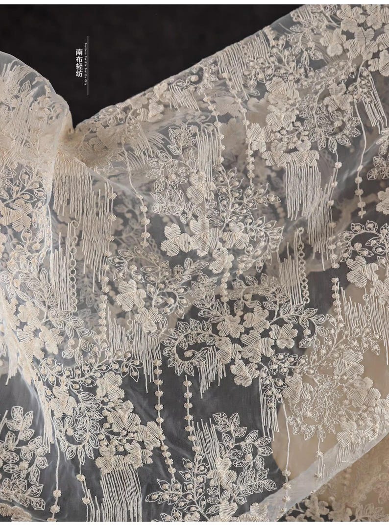Peut inclure: Tissu blanc transparent avec un motif d&eacute;licat de broderie florale et des d&eacute;tails de franges. Le tissu a une texture douce et fluide, id&eacute;al pour la cr&eacute;ation de v&ecirc;tements, d'accessoires ou de d&eacute;coration int&eacute;rieure.