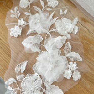 May include: White floral lace applique with 3D flowers, leaves, and pearl accents. The delicate design features various sizes of flowers and leaves, creating a textured and elegant look. Perfect for bridal wear or craft projects.