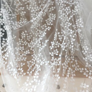 May include: White embroidered tulle fabric with a delicate floral pattern. The design features white leaves and branches on a sheer, white tulle background.  This elegant fabric is ideal for wedding apparel, bridal gowns, veils, or other special occasion garments. The embroidered floral motif adds a touch of sophistication and romance.