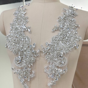 May include: Two silver rhinestone appliques with a floral design. The appliques are densely covered with sparkling rhinestones of various sizes, creating a shimmering effect. They are placed on a sheer, light-coloured fabric, likely for embellishment on garments.