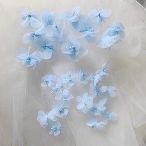 May include: A collection of light blue fabric flowers featuring delicate, translucent petals and small, round, matching blue beads at the center of each blossom. These decorative floral elements are arranged in a scattered pattern.