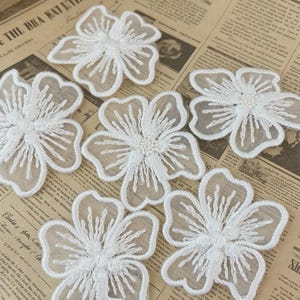 May include: White floral lace appliques. Each applique features a delicate, five-petal flower design with intricate embroidered details. The center of each flower is adorned with small, white beads. These appliques are ideal for embellishing garments or craft projects.