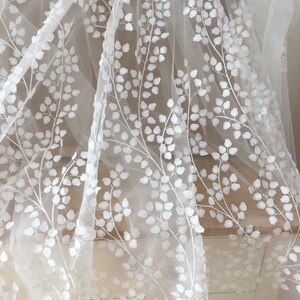 May include: White embroidered tulle fabric with a floral pattern. The fabric features delicate, raised white flowers and stems on a sheer, white tulle background.  The design is repetitive and creates an elegant, romantic look. This fabric is ideal for wedding dresses, bridal veils, and other special occasion garments.