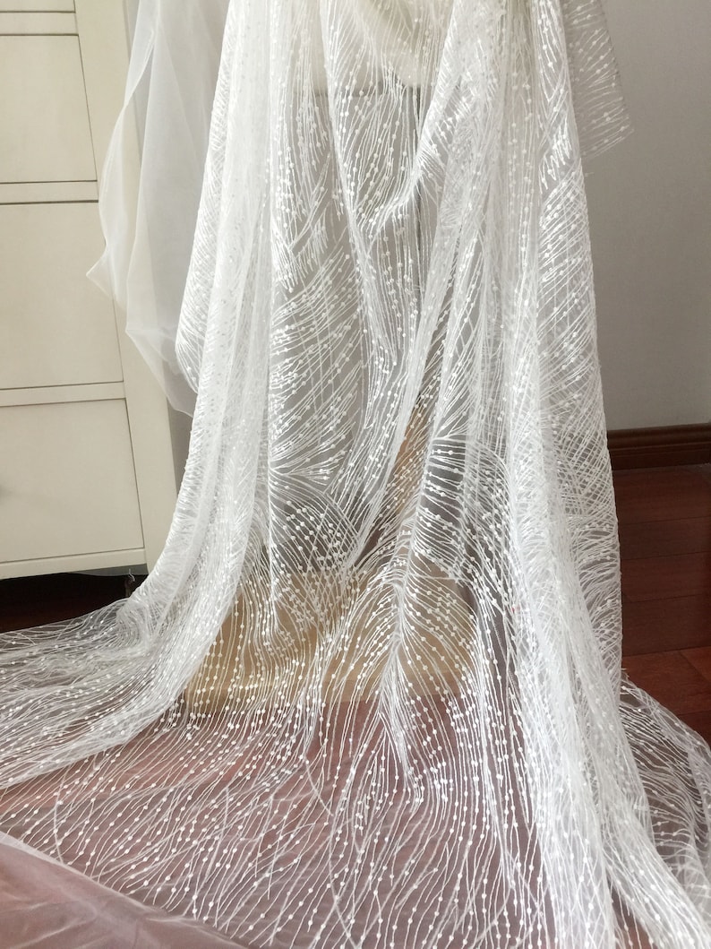 Peut inclure: Tissu de dentelle blanche perl&eacute;e avec un motif d&eacute;licat de petites perles blanches et de fines lignes. Le tissu est transparent et a une texture douce et fluide.