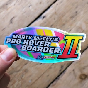 以下が含まれることがあります： 青、ピンク、黄色のカラーリングで「Marty McFly's Pro Hover Boarder II Part」というテキストが書かれたホログラフィックステッカーです。