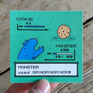 May include: A square sticker with a pixelated Cookie Monster character and a chocolate chip cookie on a green background. The text reads "COOKIE: L1" and "MONSTER: L66". The bottom text says "MONSTER used OM NOM NOM NOM!"