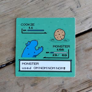 K&ouml;nnte beinhalten: Ein quadratischer, gr&uuml;ner Untersetzer mit einem pixeligen Cookie Monster und einem Schokoladen-Chip-Cookie-Design. Das Design imitiert eine Retro-Videospiel-Oberfl&auml;che mit den Texten "COOKIE", "MONSTER" und "OM NOM NOM NOM!"
