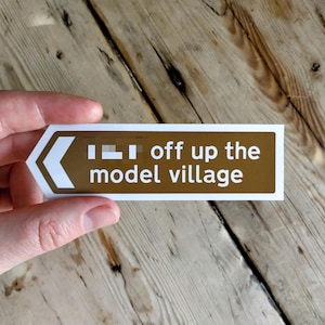 May include: A brown and white directional sign with the text "off up the model village". The sign has a white arrow pointing left. The sign is held in a hand.