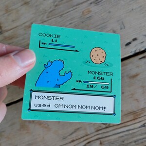 K&ouml;nnte beinhalten: Ein rechteckiger Aufkleber mit pixeligem Design, der ein blaues Monster und einen Keks zeigt. Der Text lautet "COOKIE L1 HP:" und "MONSTER 166 HP: 19/69". Darunter steht "MONSTER used OM NOM NOM NOM!" auf gr&uuml;nem Hintergrund.