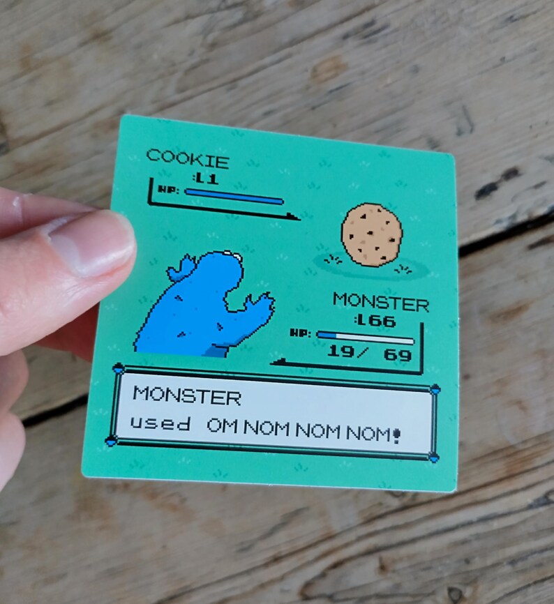 K&ouml;nnte beinhalten: Ein rechteckiger Aufkleber mit pixeligem Design, der ein blaues Monster und einen Keks zeigt. Der Text lautet "COOKIE L1 HP:" und "MONSTER 166 HP: 19/69". Darunter steht "MONSTER used OM NOM NOM NOM!" auf gr&uuml;nem Hintergrund.