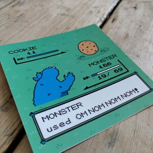 K&ouml;nnte beinhalten: Eine gr&uuml;ne Karte mit einem pixeligen Cookie Monster und einem Keks. Die Karte enth&auml;lt Text mit der Aufschrift "COOKIE" und "MONSTER" mit Gesundheitsbalken und dem Satz "MONSTER used OM NOM NOM!"