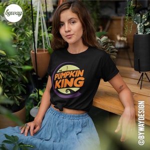 Puede incluir: Una camiseta negra con un dise&ntilde;o gr&aacute;fico morado y naranja que dice "The Pumpkin King".