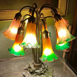 Könnte beinhalten: Dekorative Tischlampe mit sechs blütenförmigen Schirmen. Die Farbtöne gehen von Orange nach Grün über, mit einem gerüschten Rand. Der Sockel ist bronzefarben und zeigt Seerosenblätter. Die Lampe ist beleuchtet und strahlt ein warmes Licht aus.