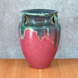 May include: A handcrafted ceramic vase with a unique design. The vase features a vibrant pink base, transitioning to a dark teal and brown top with green accents. It has two eye-like openings and a textured, dripped glaze effect.