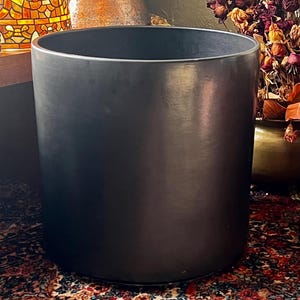 May include: A large, cylindrical black planter with a smooth, matte finish. The planter has a wide opening and a simple, modern design. It is sitting on a patterned rug, with other decorative items in the background.