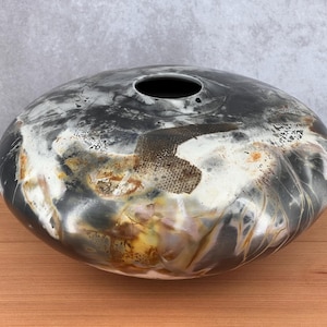 May include: A handmade ceramic vase with an unusual, rounded form. The vessel displays a dark, marbled finish with tones of black, white, and touches of gold and brown. It has a single aperture at the top and sits on a wooden surface.