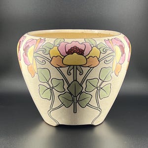 May include: A ceramic planter with a cream-coloured base featuring a crackle glaze finish. The exterior is decorated with a repeating floral pattern in shades of pink, yellow, and green, showcasing stylised flowers and leaves with dark outlines.